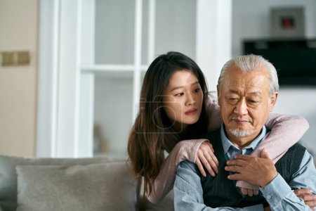Foto de Sad senior asian father sitting on couch in living room at home consoled by adult daughter - Imagen libre de derechos