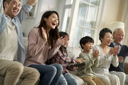 Téléchargez les photos : Multi generational asian family sitting on couch in living room at home watching sports game on tv - en image libre de droit