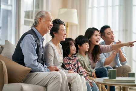 Foto de Tres generación asiático familia sentado en sofá en casa viendo tv juntos feliz y sonriente - Imagen libre de derechos