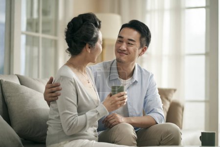 Photo for Asian adult son sitting on couch at home chatting conversing, with senior mother happy and smiling - Royalty Free Image