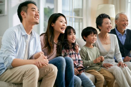 Foto de Tres generación asiático familia sentado en sofá en casa viendo tv juntos feliz y sonriente - Imagen libre de derechos