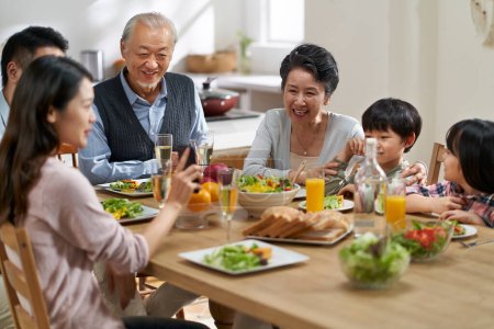 Téléchargez les photos : Young asian woman sharing pictures in cellphone with three generation family while eating meal together - en image libre de droit