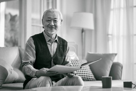 Téléchargez les photos : Happy asian old man sitting on couch at home reading a book looking at camera smiling, black and white - en image libre de droit