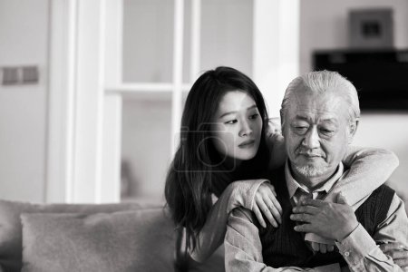 Téléchargez les photos : Triste père asiatique senior assis sur le canapé dans le salon à la maison consolé par fille adulte, noir et blanc - en image libre de droit