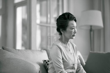 Téléchargez les photos : Vue latérale de triste femme asiatique âgée assise seule sur le canapé dans le salon à la maison, noir et blanc - en image libre de droit