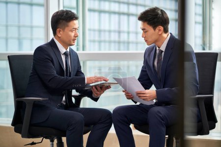 Téléchargez les photos : Deux asiatiques hommes d'affaires assis dans des chaises par la fenêtre discuter affaires au bureau - en image libre de droit
