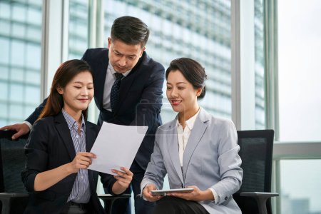 Photo for Team of three asian businesspeople sitting standing by window discussing business in office - Royalty Free Image