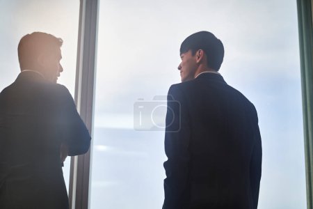 Photo for Two asian business people colleage standing by the window talking chatting discussing while looking at city view in office - Royalty Free Image