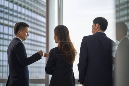 Photo for Team of three asian business people colleagues teammates standing by the window in office bumping fists - Royalty Free Image