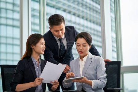 Photo for Team of three asian businesspeople sitting standing by window discussing business in office - Royalty Free Image