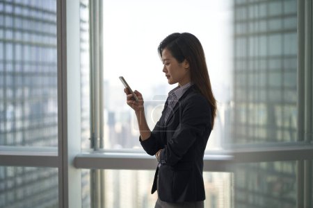 Photo for Young asian business woman standing by the window looking at cellphone in modern office - Royalty Free Image