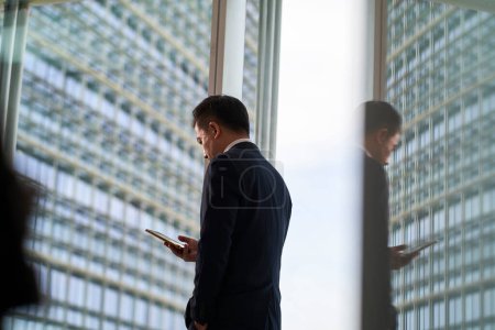 Photo for Asian businessman looking at mobile phone in office with modern building in background and in glass reflection - Royalty Free Image