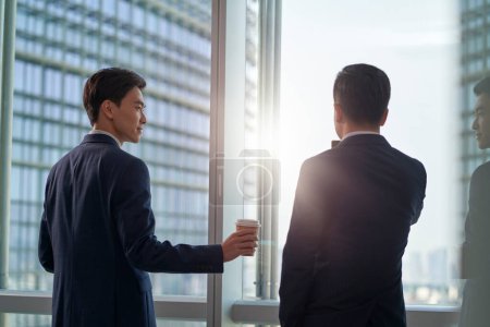 Photo for Rear view of young asian businessman handing a cup of coffee to colleague coworker teammate in front of window in modern office - Royalty Free Image
