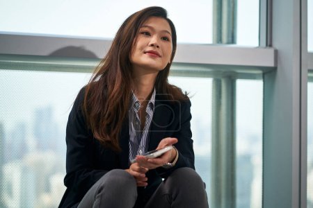 Photo for Young asian business woman sitting on the window sill taking a break in modern office - Royalty Free Image
