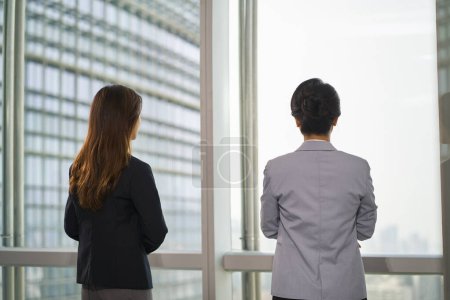 Photo for Rear view of asian business woman standing by the window in modern office looking at city view thinking - Royalty Free Image