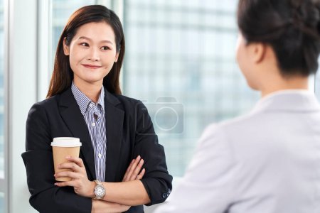 Photo for Two young asian business women standing chatting by window in modern office - Royalty Free Image