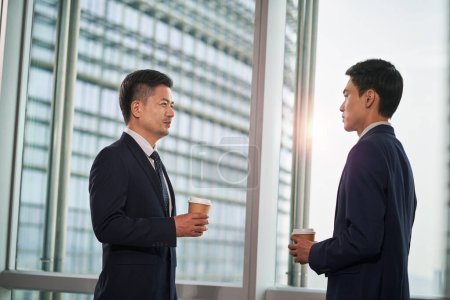 Foto de Dos asiático hombres de negocios de pie por la ventana en moderno oficina tener un chat conversación, vista lateral - Imagen libre de derechos
