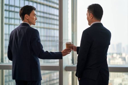Photo for Rear view of young asian businessman handing a cup of coffee to colleague coworker teammate in front of window in modern office - Royalty Free Image