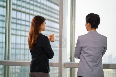 Photo for Rear view of two asian businesswomen standing by the window in modern office having a chat discussion conversation - Royalty Free Image
