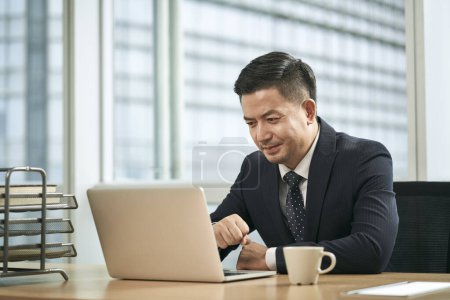 Photo for Mid adult asian businessman sitting at desk working in office using laptop computer - Royalty Free Image