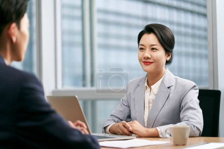 Photo for Young asian businesswoman female manager sitting at desk talking to male colleague in modern office - Royalty Free Image