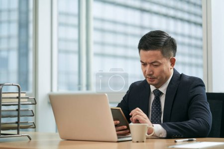 Foto de Mediados de adulto asiático hombre de negocios sentado en el escritorio de trabajo en la oficina utilizando ordenador portátil - Imagen libre de derechos