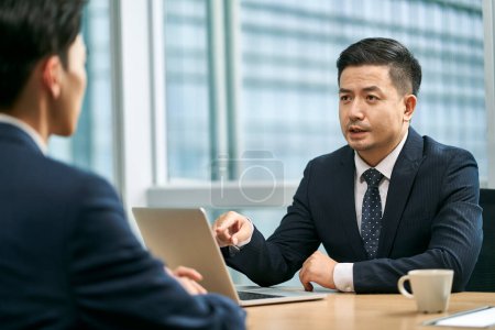 Photo for Two asian business men sitting at desk face to face in modern office having a discussion - Royalty Free Image