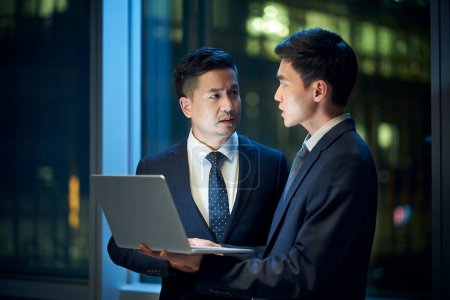 Photo for Two asian corporate executives standing by the window in office discussing business using laptop computer at night - Royalty Free Image