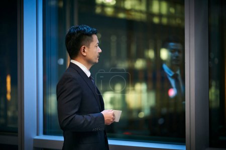 Photo for Asian corporate executives standing by the window thinking looking at night scene with a cup of coffee in hand - Royalty Free Image