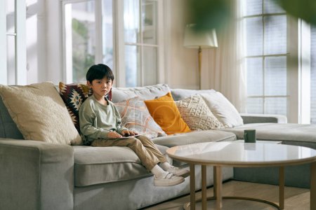 Photo for Little asian boy sitting on family couch in living room at home looking sad - Royalty Free Image