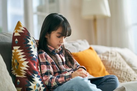 Photo for Seven-year-old little asian girl sitting on family couch at home using digital tablet computer - Royalty Free Image