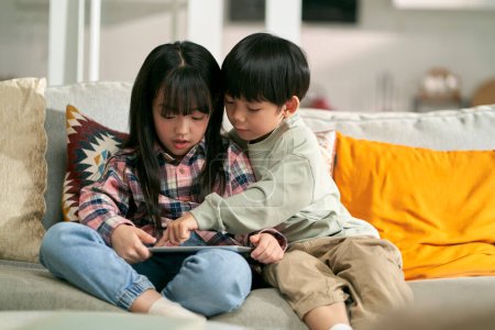 Photo for Little asian children brother and sister sitting on family couch at home using digital tablet computer together - Royalty Free Image