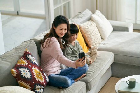 Téléchargez les photos : Heureux asiatique mère et fils assis sur la famille canapé à la maison prendre un selfie à l'aide de téléphone mobile - en image libre de droit
