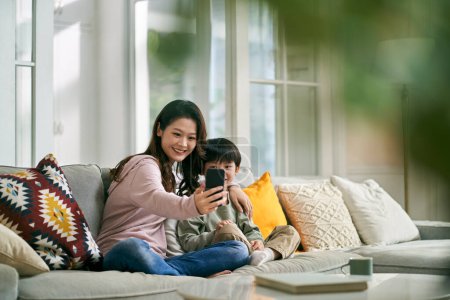 Foto de Feliz asiático madre y hijo sentado en familia sofá en casa tomando un selfie usando móvil teléfono - Imagen libre de derechos