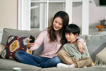 Foto de Feliz asiático madre y hijo sentado en familia sofá en casa tomando un selfie usando móvil teléfono - Imagen libre de derechos