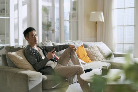 Photo for Young asian adult man sitting on family couch in living room at home looking at cellphone - Royalty Free Image