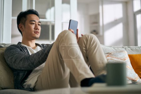 Téléchargez les photos : Jeune homme adulte asiatique assis sur le canapé familial dans le salon à la maison en regardant le téléphone portable - en image libre de droit