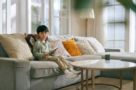 Téléchargez les photos : Petit asiatique garçon assis sur la famille canapé dans salon à la maison en regardant caméra - en image libre de droit