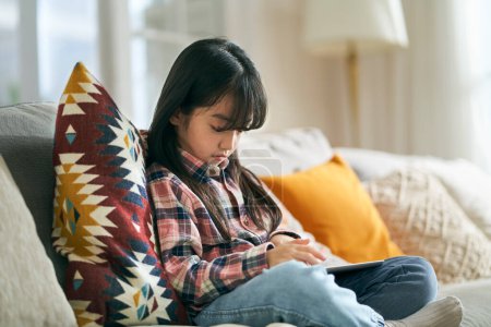 Photo for Seven-year-old little asian girl sitting on family couch at home using digital tablet computer - Royalty Free Image