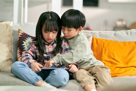 Foto de Dos asiático niños hermano y hermana sentado en familia sofá en casa jugando ordenador juego usando digital tablet - Imagen libre de derechos