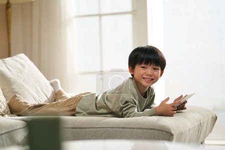 Téléchargez les photos : Mignon asiatique petit garçon couché sur l avant sur la famille canapé à la maison tenant une tablette numérique - en image libre de droit