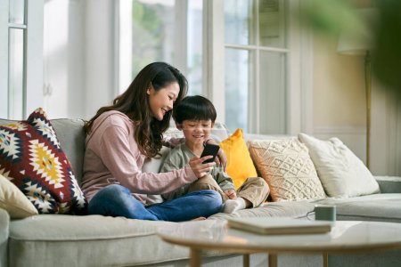 Photo for Happy asian mother and son sitting on family couch looking at cellphone photos together - Royalty Free Image