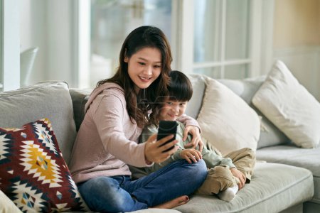 Photo for Happy asian mother and son sitting on family couch at home taking a selfie using mobile phone - Royalty Free Image