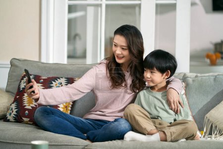 Photo for Happy asian mother and son sitting on family couch at home taking a selfie using mobile phone - Royalty Free Image