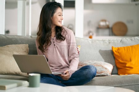 Photo for Happy young asian business woman sitting on couch at home working using laptop computer - Royalty Free Image