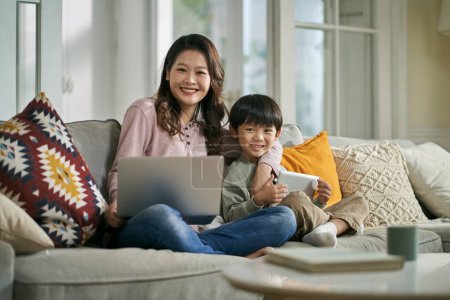 Photo for Beautiful asian mother and five-year-old son sitting on family couch at home looking at camera smiling - Royalty Free Image