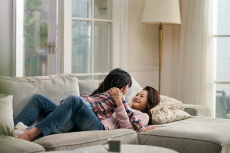 Téléchargez les photos : Jeune asiatique mère couché sur la famille canapé avoir un face à face agréable conversation avec sept ans fille - en image libre de droit