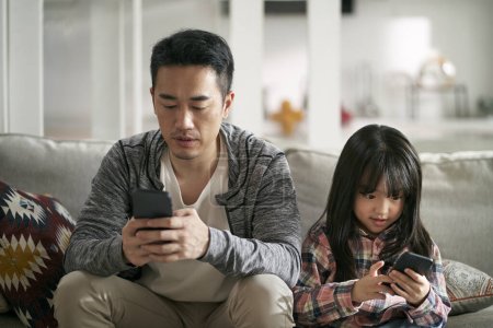 Téléchargez les photos : Jeune asiatique père et sept ans fille assis sur la famille canapé en utilisant téléphone cellulaire ensemble regarder grave - en image libre de droit