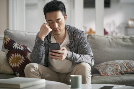 Téléchargez les photos : Jeune asiatique homme assis sur canapé à la maison en regardant le téléphone cellulaire à la recherche grave - en image libre de droit
