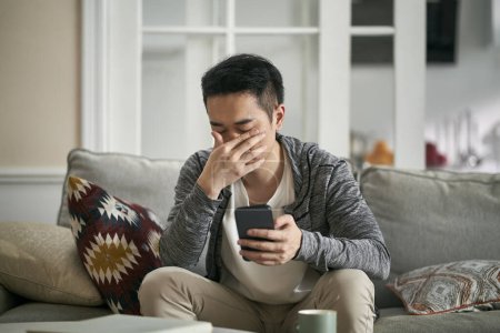 Photo for Young asian sitting on couch at home covering eyes with hand feeling tired after looking at cellphone for long time - Royalty Free Image